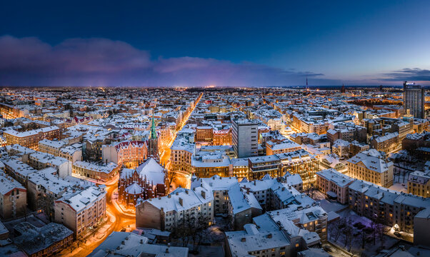 Riga city panorama in winter time. Street lights glowing in blue hour. Snowy rooftops. Iconic buildings and architecture. Drone footage. 