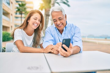 Middle age couple in love sitting at the terrace of coffee shop using smartphone happy and cheerful together