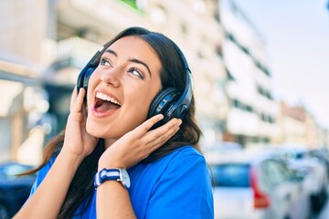 Young hispanic woman smiling happy listening to music using headphones walking at the city.