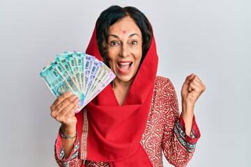 Middle age hispanic woman wearing traditional indian clothes and holding rupee banknotes screaming proud, celebrating victory and success very excited with raised arm