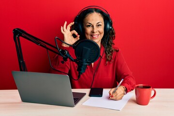 Beautiful middle age woman working at radio studio doing ok sign with fingers, smiling friendly gesturing excellent symbol