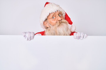 Old senior man with grey hair and long beard wearing santa claus costume holding banner smiling looking to the side and staring away thinking.