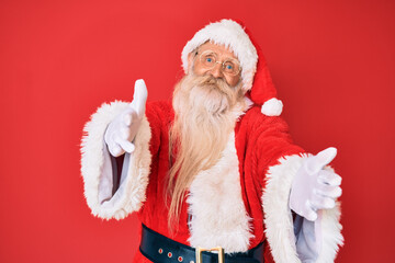 Old senior man with grey hair and long beard wearing traditional santa claus costume looking at the camera smiling with open arms for hug. cheerful expression embracing happiness.