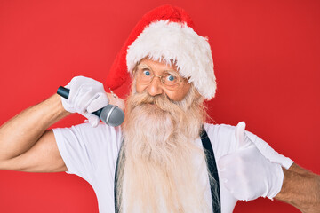 Old senior man with grey hair and long beard wearing santa claus costume singing with microphone smiling happy and positive, thumb up doing excellent and approval sign