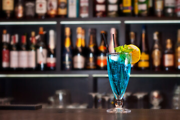 Blue Lagoon cocktail. Blue cocktail on the bar counter. Drink with martini, vodka, rum, tequila and fruit on the background of bottles with alcohol. Glass with booze on a wooden board.