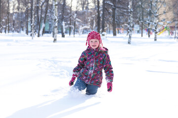 Fototapeta na wymiar Winter activities outdoors. Positive little girl wearing a warm clothes walking through the snowdrifts in snowy park on cheerful winter day