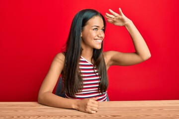 Beautiful hispanic woman wearing casual clothes sitting on the table very happy and smiling looking far away with hand over head. searching concept.