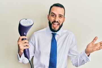 Naklejka premium Young business man with beard holding electric steam iron celebrating achievement with happy smile and winner expression with raised hand