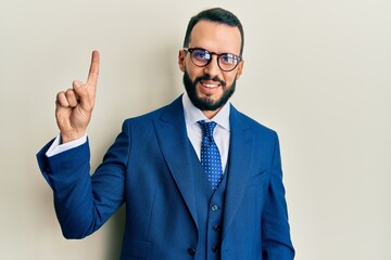 Young man with beard wearing business suit and tie showing and pointing up with finger number one while smiling confident and happy.