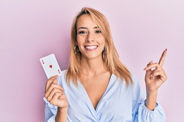 Beautiful blonde woman holding ace poker card smiling happy pointing with hand and finger to the side