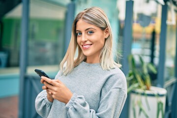 Young blonde girl smiling happy using smartphone at the city