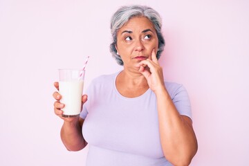 Senior hispanic woman holding glass of milk serious face thinking about question with hand on chin, thoughtful about confusing idea