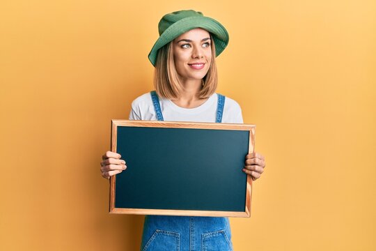 Young caucasian blonde woman holding blackboard smiling looking to the side and staring away thinking.
