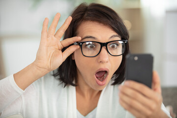 woman wearing eyeglasses looking with shock at smartphone - obrazy, fototapety, plakaty