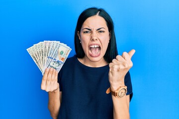 Beautiful young woman holding dollars angry and mad raising fist frustrated and furious while...