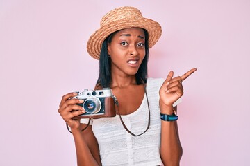 Young african american woman wearing summer hat holding vintage camera pointing aside worried and nervous with forefinger, concerned and surprised expression