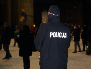 police officer at work observing protesters during evening street demonstration, in background LGBT flag
