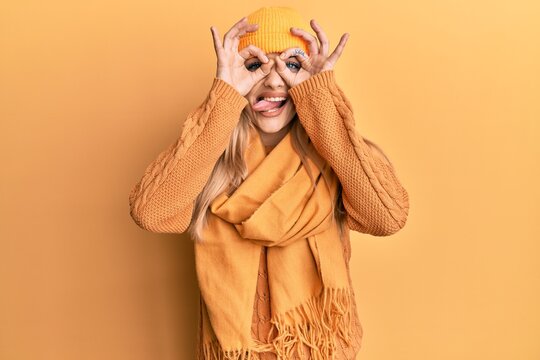 Young caucasian woman wearing wool winter sweater and cap doing ok gesture like binoculars sticking tongue out, eyes looking through fingers. crazy expression.
