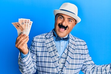 Mature middle east man with mustache wearing elegant vintage style holding pounds banknotes celebrating achievement with happy smile and winner expression with raised hand