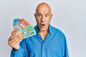 Middle age bald man holding australian dollars scared and amazed with open mouth for surprise, disbelief face