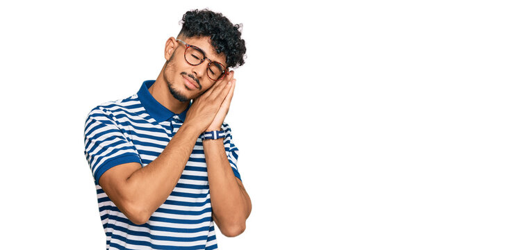 Young arab man wearing casual clothes and glasses sleeping tired dreaming and posing with hands together while smiling with closed eyes.