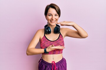 Obraz premium Young brunette woman with short hair wearing sportswear and headphones gesturing with hands showing big and large size sign, measure symbol. smiling looking at the camera. measuring concept.