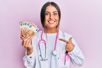 Beautiful hispanic woman wearing medical uniform holding 50 euros banknotes smiling happy pointing...