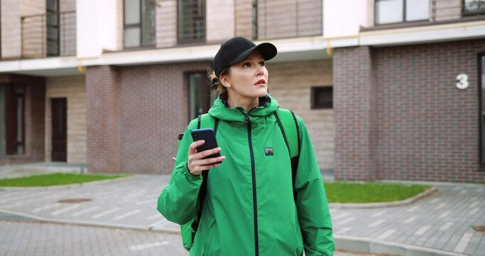 Portrait of young female employee with big backpack walk on street and texting on smartphone looking for direction to deliver orders. Caucasian delivery woman typing on cellphone searching for address