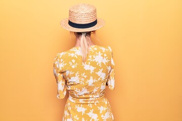 Beautiful blonde woman on vacation wearing summer hat and dress over yellow background standing...