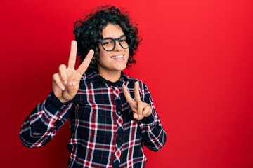 Young hispanic woman with curly hair wearing casual clothes and glasses smiling looking to the camera showing fingers doing victory sign. number two.