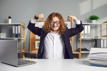 Happy office worker in eyeglasses smiling and showing motivation for work. Funny man in suit sitting at desk and flexing arm muscles. Cheerful employee feeling energetic and ready for productive day