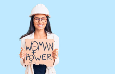 Young beautiful latin girl wearing architect hardhat holding woman power banner looking positive and happy standing and smiling with a confident smile showing teeth