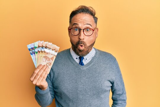 Handsome middle age business man holding canadian dollars scared and amazed with open mouth for surprise, disbelief face