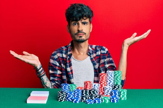 Young Hispanic Man Sitting On The Table With Poker Chips And Cards Clueless And Confused With Open Arms, No Idea And Doubtful Face.