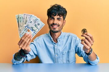 Young hispanic man holding dollars and bitcoin winking looking at the camera with sexy expression,...