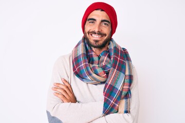 Handsome young man with curly hair and bear wearing a winter sweater, scarf and wool hat happy face smiling with crossed arms looking at the camera. positive person.