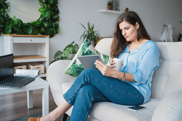 Business woman in blue shirt typing in tablet.Online conference.Remote work at home office sitting on couch.Message,dating,meeting in social net.Relax in fasionable appartment.Covid-19 self isolation