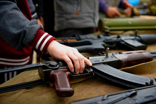 A child's hand takes an automatic rifle from the table with weapons. Close-up photo.
