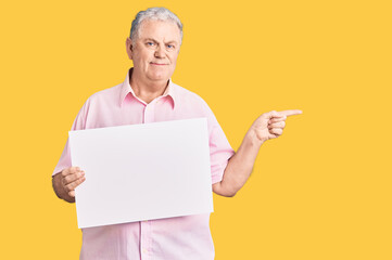 Senior grey-haired man holding blank empty banner smiling happy pointing with hand and finger to the side