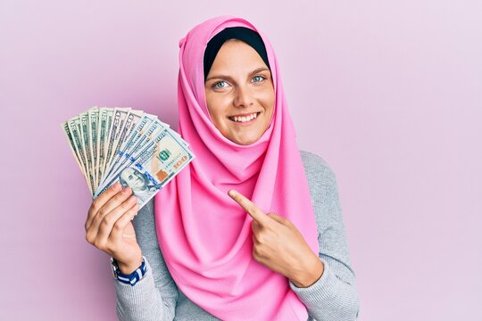 Young caucasian woman wearing islamic hijab holding dollars banknotes smiling happy pointing with hand and finger