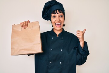 Beautiful brunettte woman professional chef holding take away food pointing thumb up to the side smiling happy with open mouth