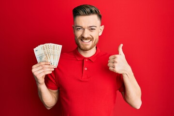 Young redhead man holding 100 danish krone banknotes smiling happy and positive, thumb up doing excellent and approval sign