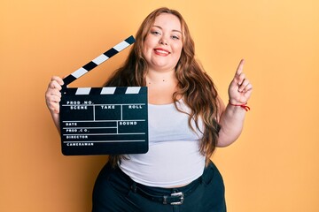 Plus size caucasian young woman holding video film clapboard smiling happy pointing with hand and...