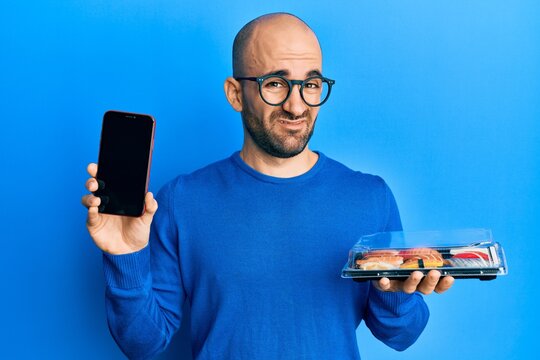 Young hispanic man holding take away food showing smartphone screen clueless and confused expression. doubt concept.