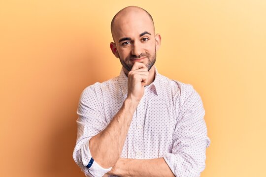 Young handsome bald man wearing elegant shirt smiling looking confident at the camera with crossed arms and hand on chin. thinking positive.
