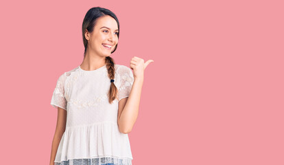Young beautiful girl wearing casual t shirt smiling with happy face looking and pointing to the side with thumb up.