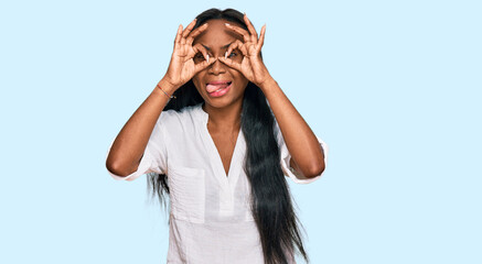 Young black woman wearing casual clothes doing ok gesture like binoculars sticking tongue out, eyes looking through fingers. crazy expression.