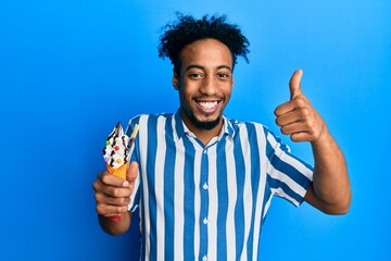 Young african american man with beard eating ice cream smiling happy and positive, thumb up doing...