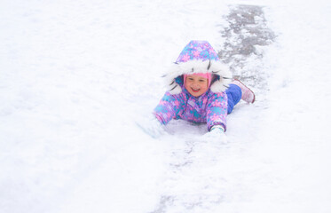 A little pretty girl in a winter overalls is riding on ice slides in winter, a child is driving...