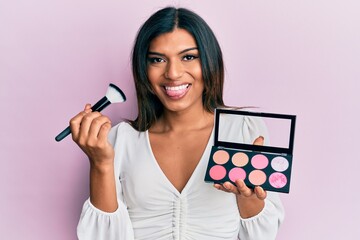 Young latin transsexual transgender woman holding makeup brush and blush palette sticking tongue out happy with funny expression.